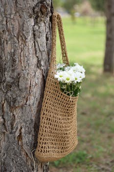 A handmade jute bag filled with white daisies hangs on a tree, showcasing eco-friendly style.